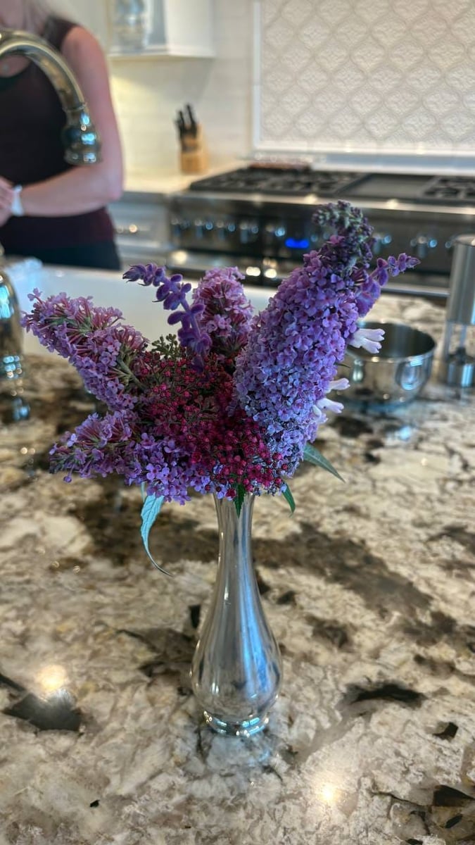 Bouquet of purple lilac and butterfly bush in a silver bud vase on a granite countertop.