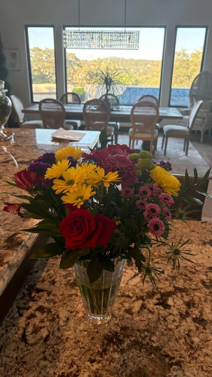 Mixed bouquet with roses, yellow daisies, mums, and greenery in a crystal vase on a granite island.